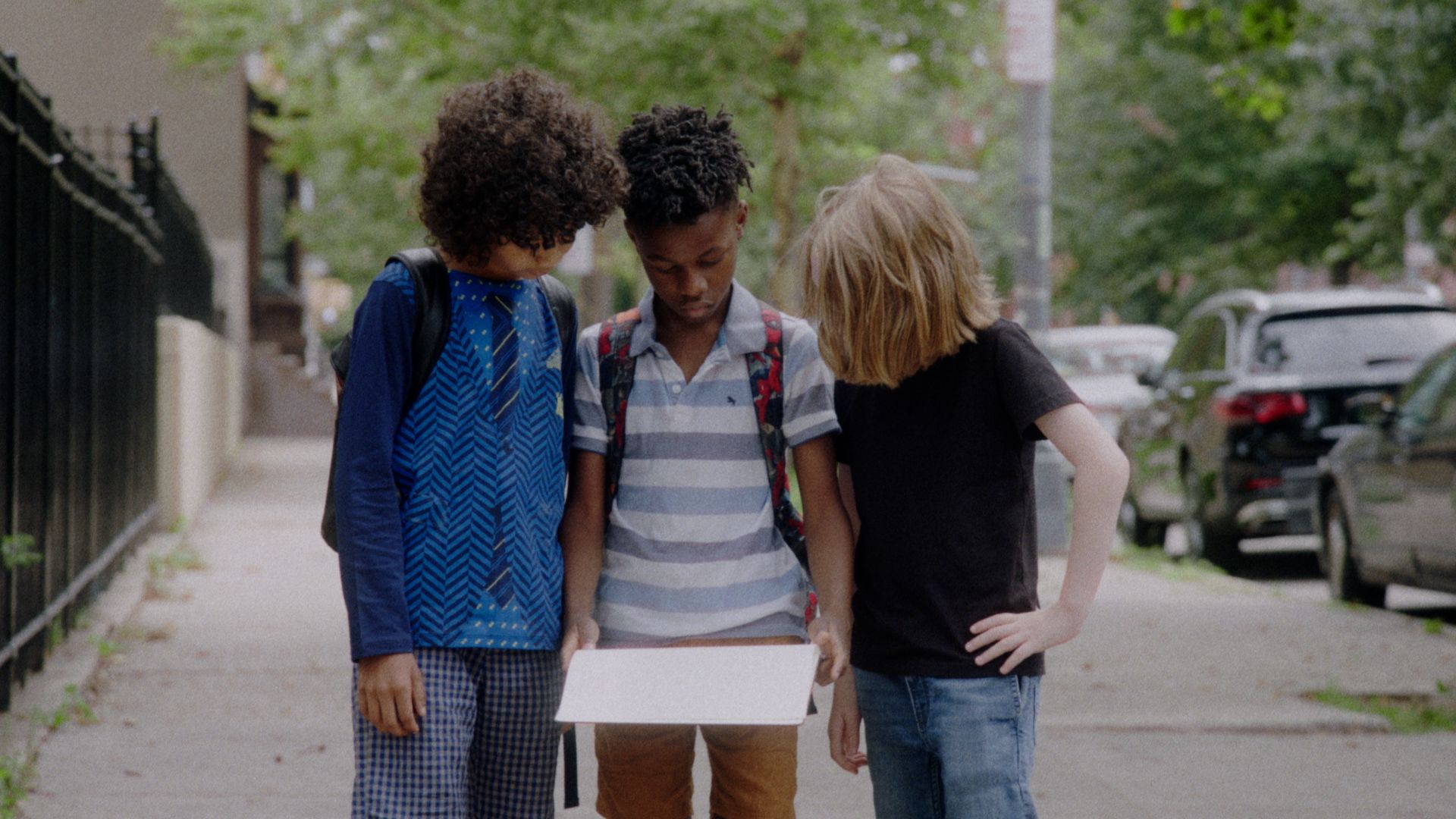 Three boys on a tree-lined sidewalk look down at a sheet of paper.