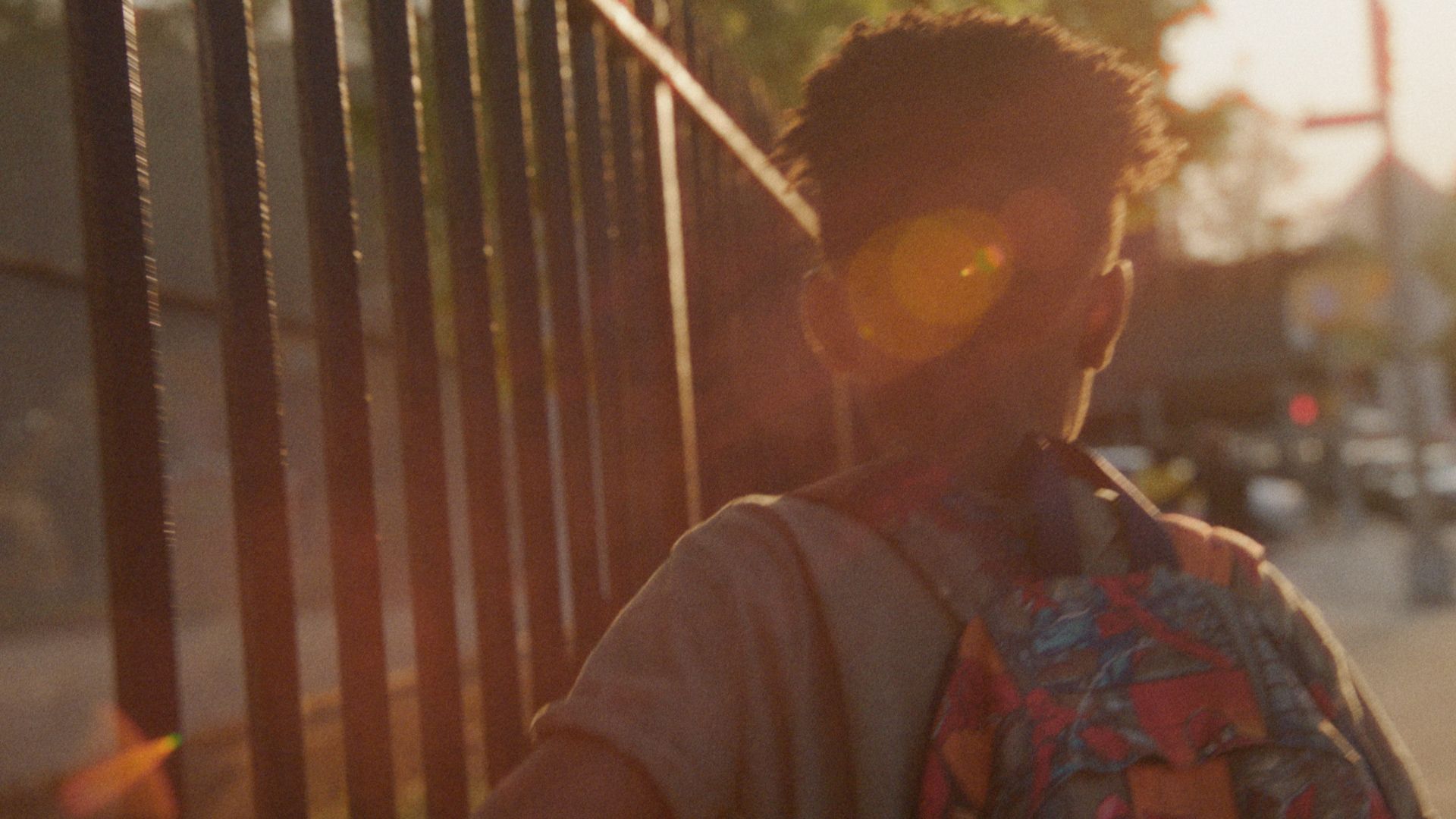 A boy with a backpack walks past an iron fence in low sun.